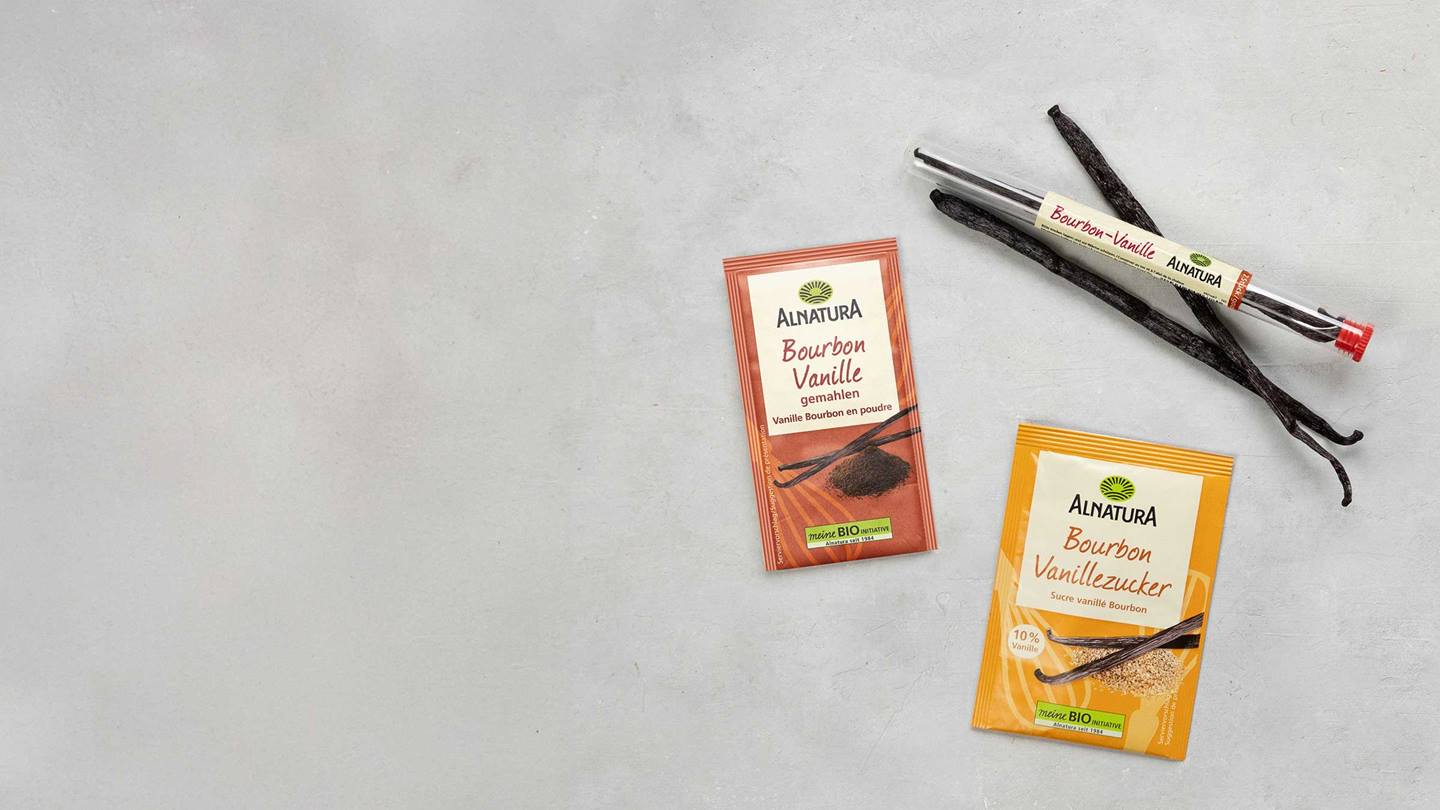 Das Bild zeigt eine Nahaufnahme von Bourbon-Vanille Produkten der Marke Alnatura auf einer neutralen Oberfläche. Zu sehen sind eine Packung gemahlene Bourbon-Vanille, eine Packung Bourbon-Vanillezucker und ein Röhrchen mit ganzen Bourbon-Vanilleschoten. Die Farben der Verpackungen sind in warmen Erdtönen gehalten.